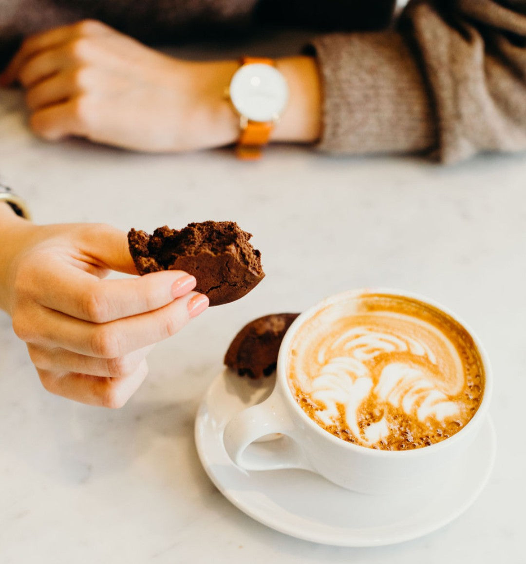 A person holds half a vegan cookie beside a cup of coffee on a table.